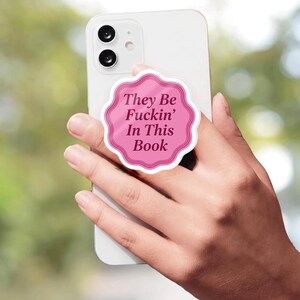 Könnte beinhalten: Ein weißes Smartphone mit einem runden, wellenförmigen Handygriff auf der Rückseite. Der Griff ist rosa mit einem dunkelrosa Rand und zeigt den schwarzen Text "They Be Fuckin' In This Book", der mittig in einer Serifenschrift gedruckt ist.