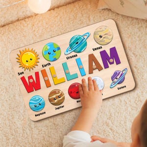 May include: Wooden puzzle featuring the name "WILLIAM" in colorful letters, with planet-shaped pieces. The planets include the Sun, Earth, Uranus, Venus, Saturn, Neptune, Jupiter, and Mars. A child's hand is interacting with the puzzle.