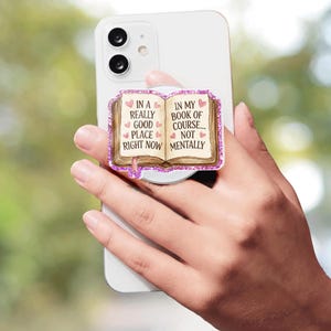 Könnte beinhalten: Ein weißes Smartphone mit einem runden Handygriff, der ein offenes Buch zeigt. Auf den Buchseiten steht der Text: IN A REALLY GOOD PLACE RIGHT NOW IN MY BOOK OF COURSE... NOT MENTALLY, verziert mit kleinen rosa Herzen und glitzernden Rändern.