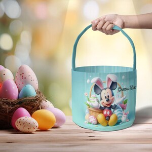 May include: A light blue Easter basket featuring Mickey Mouse with bunny ears, surrounded by Easter eggs. The basket has a handle and the words "Custom Text". A nest of colorful Easter eggs sits on a wooden surface.