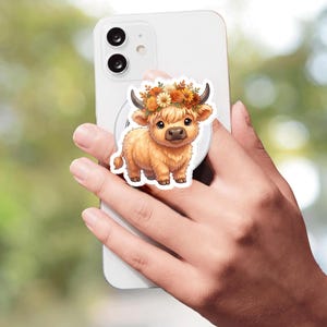 May include: A white smartphone featuring a circular phone grip on the back. The grip displays a cartoon illustration of a fluffy light brown Highland cow with a floral crown of orange, yellow, and brown flowers on its head, set against a white background.
