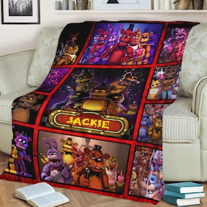 May include: A colorful fleece blanket featuring a collage of characters from the Five Nights at Freddy's video game franchise. The blanket is divided into squares with different characters in each square. The blanket has a red border and the name "JACKIE" is written in a marquee sign at the top of the blanket.