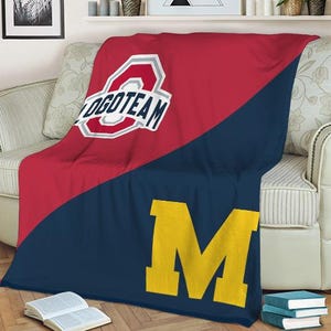 May include: A red and navy blue blanket with a diagonal color block design. The top half is red with a white and gray logo that says "LOGOTEAM". The bottom half is navy blue with a large yellow "M".