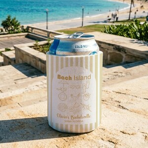 Op de afbeelding: Een drankblikkoeler met verticale beige en witte strepen. De voorzijde toont de tekst Bach Island, Olivia's Bachelorette en Miami Florida, vergezeld van goudkleurige lijntekeningen van strandartikelen zoals schelpen en sandalen.
