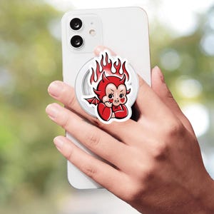 May include: A white smartphone featuring a circular collapsible grip attached to the back. The grip displays a cartoon illustration of a red devil character with small wings and flames rising from its head, set against a white background.