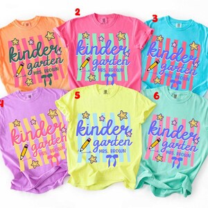 Könnte beinhalten: Sechs kurzärmelige T-Shirts in Orange, Pink, Blau, Lila, Gelb und Mintgrün. Jedes T-Shirt zeigt eine Grafik mit vertikalen Streifen, dem Text kindergarten, einem Bleistift, Sternen, einer Schleife und dem personalisierten Text MRS. BROWN.