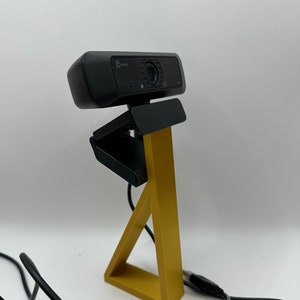 May include: A black webcam with a gold triangular stand. The webcam has a microphone and a lens cover. The stand is made of plastic and has a textured surface.