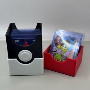 May include: A black and white card storage box designed like a Poké Ball, holding a stack of trading cards in protective sleeves, next to a separate red card storage box containing a stack of trading cards featuring a Spongebob Squarepants graphic.