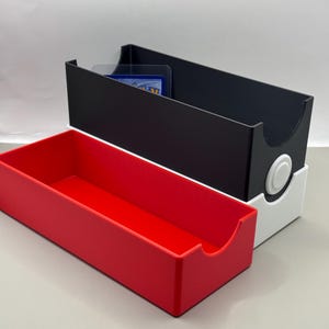 May include: A rectangular storage box for trading cards featuring a black upper section, a white middle band with a circular white button detail, and a bright red lower tray. The box has curved cutouts on the sides for easy access to the contents.