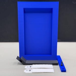 May include: A vibrant blue picture frame with a rectangular design. The frame is accompanied by a black support piece, two white adhesive strips, and a blue mounting bracket with a screw. The frame is designed to hold a picture or artwork.