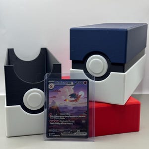 May include: A Charizard ex trading card in a clear protective sleeve positioned in front of two rectangular storage boxes. One box is blue and white with a circular button, and the other is black and white with a circular button, both resembling Poké Balls.