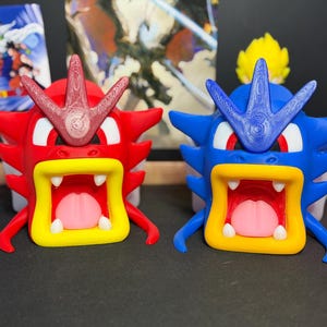 May include: Two stylized creature figurines featuring wide-open mouths with yellow borders, white teeth, and pink tongues. One figurine is primarily red with a textured crest, while the other is blue with a matching textured crest and red eyes.