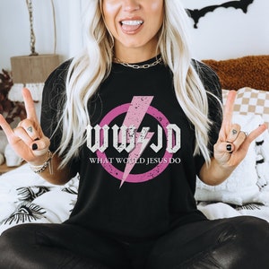 May include: Black t-shirt featuring a pink circle with a lightning bolt and the text "WWJD WHAT WOULD JESUS DO" in a distressed white font. The shirt is a casual style.