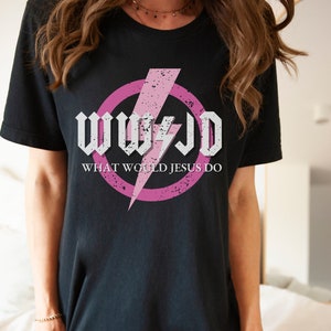 May include: Black t-shirt featuring the text "WWJD" in a white, gothic-style font, with a pink lightning bolt and a pink circle. Below the text is "WHAT WOULD JESUS DO" in a sans-serif font.