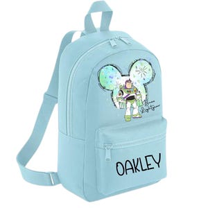 May include: Light blue backpack with a Mickey Mouse ear design. The backpack features a colourful illustration of Buzz Lightyear from Toy Story. The text "Buzz Lightyear" and the name "OAKLEY" are printed on the backpack.