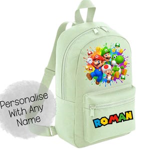 Puede incluir: Una mochila verde claro con un gráfico colorido de Mario, Luigi, Yoshi y Toad. La mochila tiene un bolsillo frontal con el nombre "ROMAN" en letras coloridas. También es visible el texto "Personalise With Any Name".