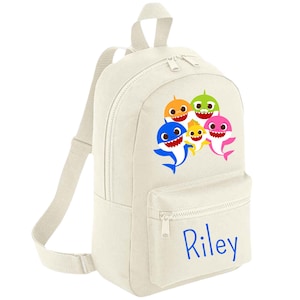 May include: A white canvas backpack featuring a colourful Baby Shark family graphic and the name "Riley" printed in blue on the front pocket.