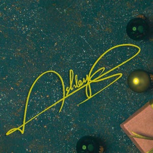 Op de afbeelding: Een teal achtergrond met een gele handtekening met de tekst "Ashley". De afbeelding bevat ook groene kerstballen en een klein ingepakt cadeau met een gouden lint, wat een feestelijk vakantiethema creëert.