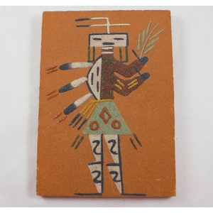May include: A rectangular sand painting featuring a stylized Native American figure. The figure is depicted with a brown torso, a green skirt, and white legs. It holds a green leaf and has multiple arms with black and white details. The background is a warm orange.