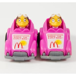 Puede incluir: Dos coches de juguete rosas con un personaje de pájaro amarillo conduciendo cada coche. Los coches tienen el texto "Birdie, the Early Bird" y el logotipo de McDonald's en el lateral.