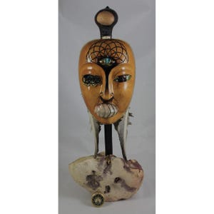 Könnte beinhalten: Handgefertigte Holzmasken-Skulptur mit hellbraunem Finish. Die Maske weist aufwendige Details auf, darunter eingelegte Augen, ein drittes Auge und ein geometrisches Muster auf der Stirn. Sie ist auf einem Steinsockel mit Federn und einem kleinen Medaillon montiert.