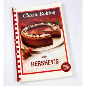 May include: A cookbook titled "Classic Baking with Hershey's" featuring a chocolate cheesecake with whipped cream. The cover has a red border and a spiral binding. Red roses are in the background. A sticker on the cover says "Valuable Hershey's Coupons Inside!"