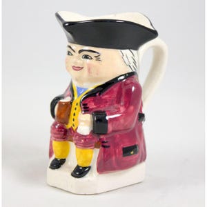 Könnte beinhalten: Keramikkrug in Form eines sitzenden Mannes, der einen schwarzen Hut und einen weinroten Mantel trägt. Die Figur hält einen Krug und ist mit gelben Kniehosen und schwarzen Schuhen bekleidet. Der Krug hat einen weißen Henkel und Ausguss.