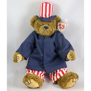 Puede incluir: Un oso de peluche marrón que lleva una chaqueta azul y pantalones y sombrero a rayas rojas y blancas. El oso lleva una etiqueta Ty que dice "Beanie Babies".