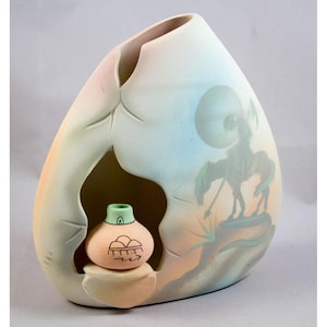 May include: A ceramic vase with a unique, egg-like shape. The vase features a cutout revealing a smaller, decorated pot inside. The exterior is painted with a Native American warrior on horseback and a landscape scene. The color palette includes soft pastels.