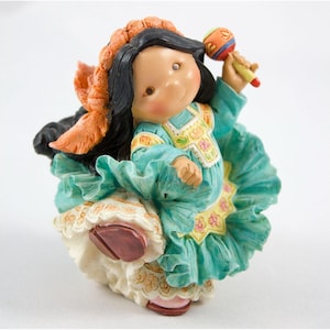May include: A figurine of a girl in a teal dress with a ruffled skirt, holding a maraca. The figurine has black hair, a floral headpiece, and a smiling expression. The dress has yellow and pink floral accents.