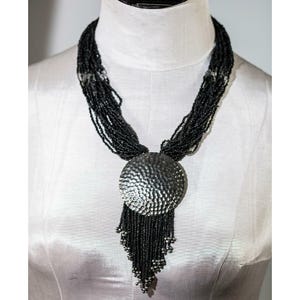 Può includere: Una collana di perline nere con un grande ciondolo argentato strutturato. Il ciondolo ha un aspetto di metallo martellato ed è rotondo. La collana è esposta su un manichino bianco.