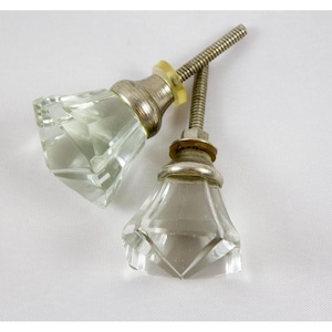 May include: Two clear glass cabinet knobs with faceted diamond-shaped designs. Each knob has a silver-colored metal base and a threaded rod for installation. The knobs are set against a white background.