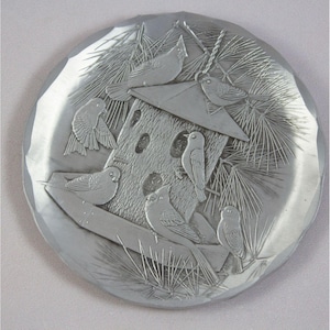 May include: A round, silver-colored decorative plate with an embossed design of a birdhouse and several birds. The birds are perched on the birdhouse, a branch, and the plate's edge. Pine needles are also embossed around the birdhouse.