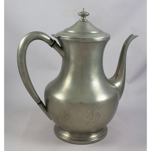 May include: A vintage silver-colored metal coffee pot with a curved handle and a spout. The pot has a lid with a decorative finial and an engraved initial on the body. The pot sits on a round base and has a classic, elegant design.