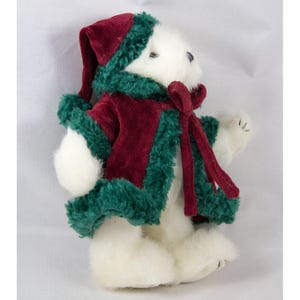 May include: A white plush teddy bear wearing a burgundy velvet coat and hat trimmed with green faux fur. A matching burgundy scarf is tied around the bear's neck. The bear's paws and face are visible.