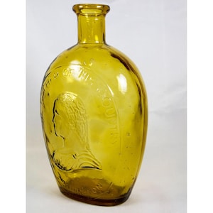 May include: A vintage, amber-colored glass bottle with an oval shape. The bottle features an embossed profile of a person and text that reads "OF THIS COUNTY" and "INGTON". The bottle has a long neck and a rounded base.
