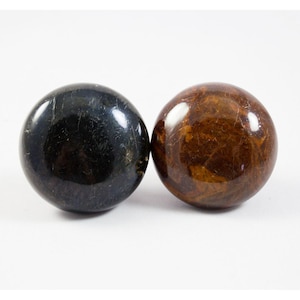 Op de afbeelding: Twee gladde, ronde stenen. Eén is zwart met een subtiele glans, de andere is bruin met een marmerpatroon.