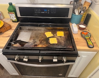 May include: A stainless steel gas range with a large rectangular griddle plate centered on the burners. Three beef patties topped with melted yellow cheese are cooking on the griddle surface, accompanied by a metal spatula resting to the side.