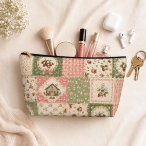 May include: A zipped pouch featuring a patchwork pattern of strawberries, small houses, and floral motifs in shades of pink, green, and cream. The pouch is filled with a makeup brush, a compact, two lip products, and a small glass perfume bottle.