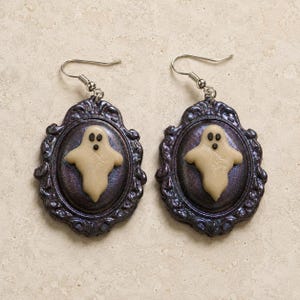 Può includere: Un paio di orecchini pendenti con una cornice ovale decorata di colore viola scuro e una figura di fantasma in rilievo color crema al centro. Gli orecchini sono fissati a ganci in metallo color argento per orecchie forate con un design dettagliato.