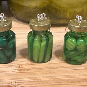 May include: Three miniature pickle jar earrings. Each jar is filled with green pickles and has a gold-colored lid. The earrings are displayed on a wooden surface, with a larger jar of pickles in the background.