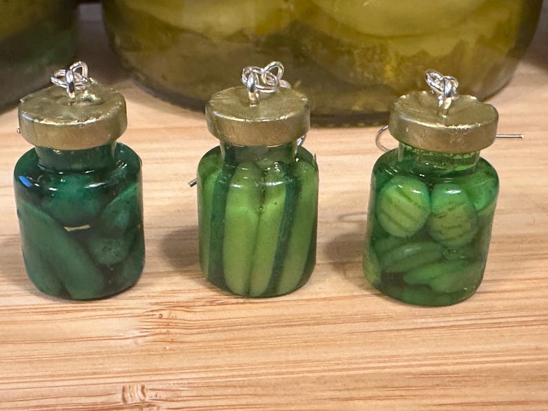 May include: Three miniature pickle jar earrings. Each jar is filled with green pickles and has a gold-colored lid. The earrings are displayed on a wooden surface, with a larger jar of pickles in the background.