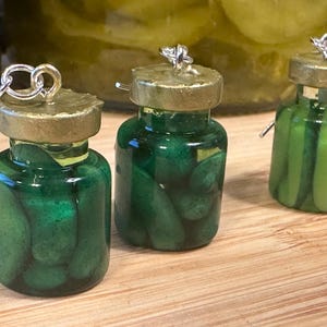 May include: Three miniature jar earrings filled with green pickles. Each jar has a gold-colored lid and a silver earring hook. The jars are displayed on a wooden surface, with a blurred background of more jars.