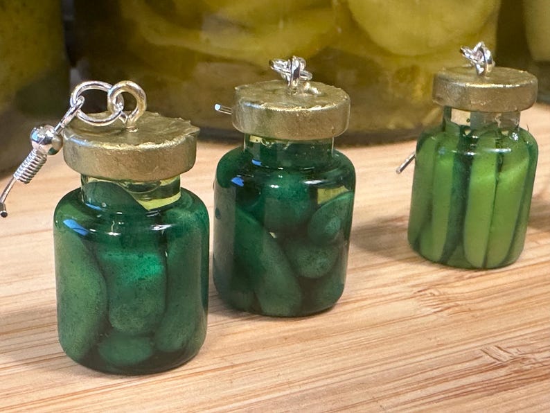 May include: Three miniature jar earrings filled with green pickles. Each jar has a gold-colored lid and a silver earring hook. The jars are displayed on a wooden surface, with a blurred background of more jars.