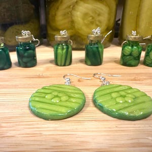 May include: A collection of handmade pickle-themed jewelry. The image features miniature glass jar pendants filled with green beads, and two green pickle slice earrings with silver hooks. The background includes jars of pickles.