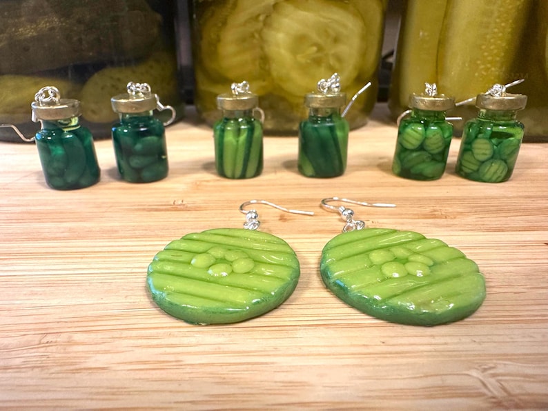 May include: A collection of handmade pickle-themed jewelry. The image features miniature glass jar pendants filled with green beads, and two green pickle slice earrings with silver hooks. The background includes jars of pickles.