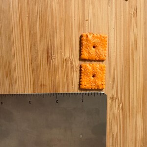 Puede incluir: Dos galletas cuadradas y naranjas con un agujero en el centro, junto a una regla met&aacute;lica. Las galletas est&aacute;n sobre una superficie de madera. La regla est&aacute; marcada con medidas en cm.