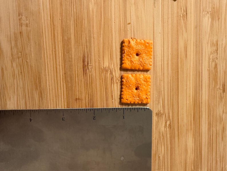 Puede incluir: Dos galletas cuadradas y naranjas con un agujero en el centro, junto a una regla met&aacute;lica. Las galletas est&aacute;n sobre una superficie de madera. La regla est&aacute; marcada con medidas en cm.