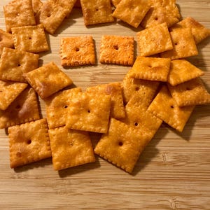 Puede incluir: Una colecci&oacute;n de galletas de queso cuadradas y naranjas dispuestas sobre una superficie de madera. Cada galleta tiene un peque&ntilde;o agujero en el centro y una superficie ligeramente texturizada. Las galletas son de color naranja dorado, lo que sugiere un aspecto horneado o tostado.