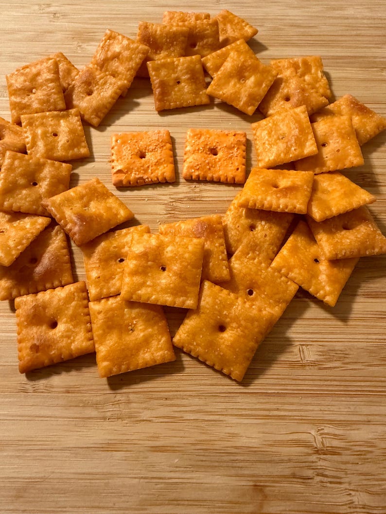 Puede incluir: Una colecci&oacute;n de galletas de queso cuadradas y naranjas dispuestas sobre una superficie de madera. Cada galleta tiene un peque&ntilde;o agujero en el centro y una superficie ligeramente texturizada. Las galletas son de color naranja dorado, lo que sugiere un aspecto horneado o tostado.